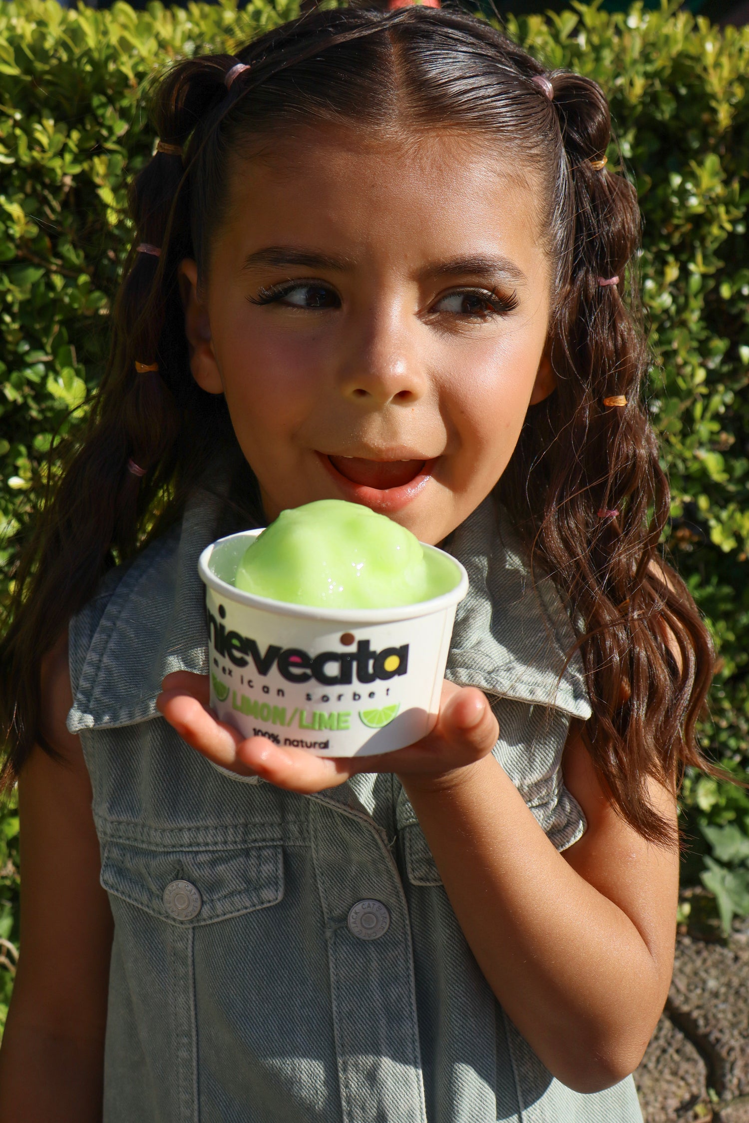 Young girl holding a cup of green lime ice cream with nievecita branding against a green hedge background.