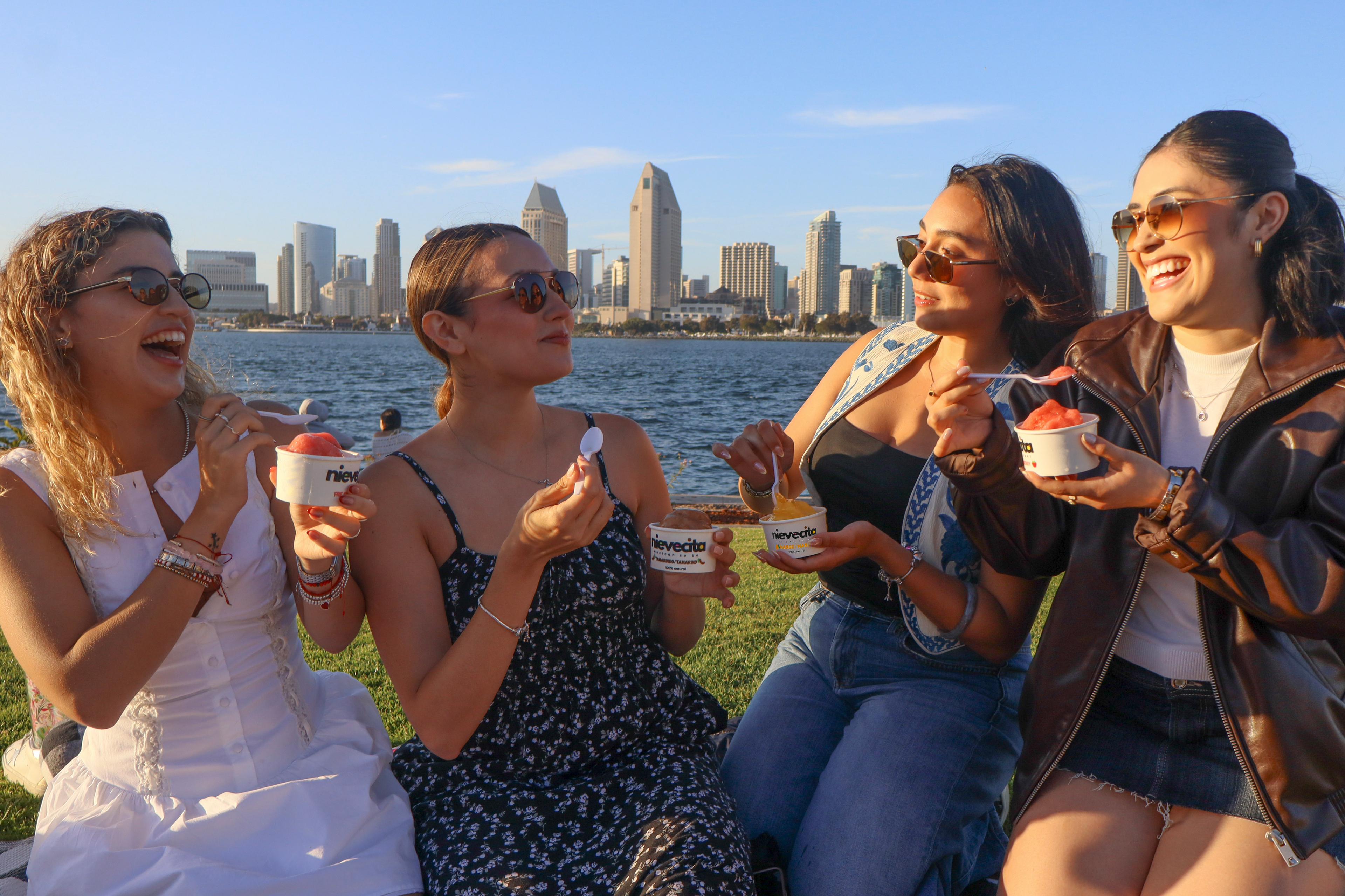 Four women enjoying ice cream sorbet by a waterfront with a city skyline in the background